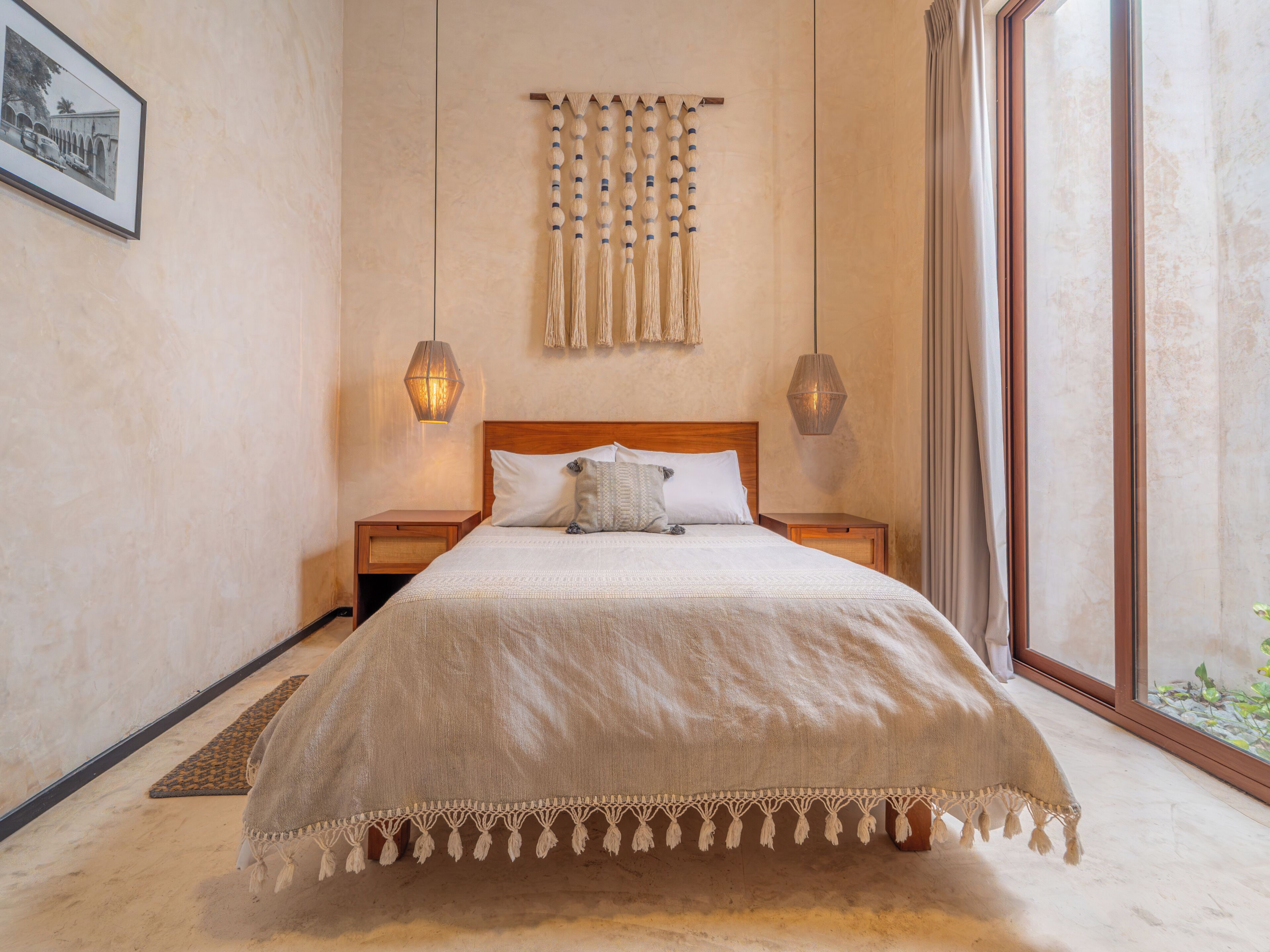 A bedroom with a large bed, wooden headboard, two bedside tables, a wall-mounted macrame piece, and a large window with curtains.