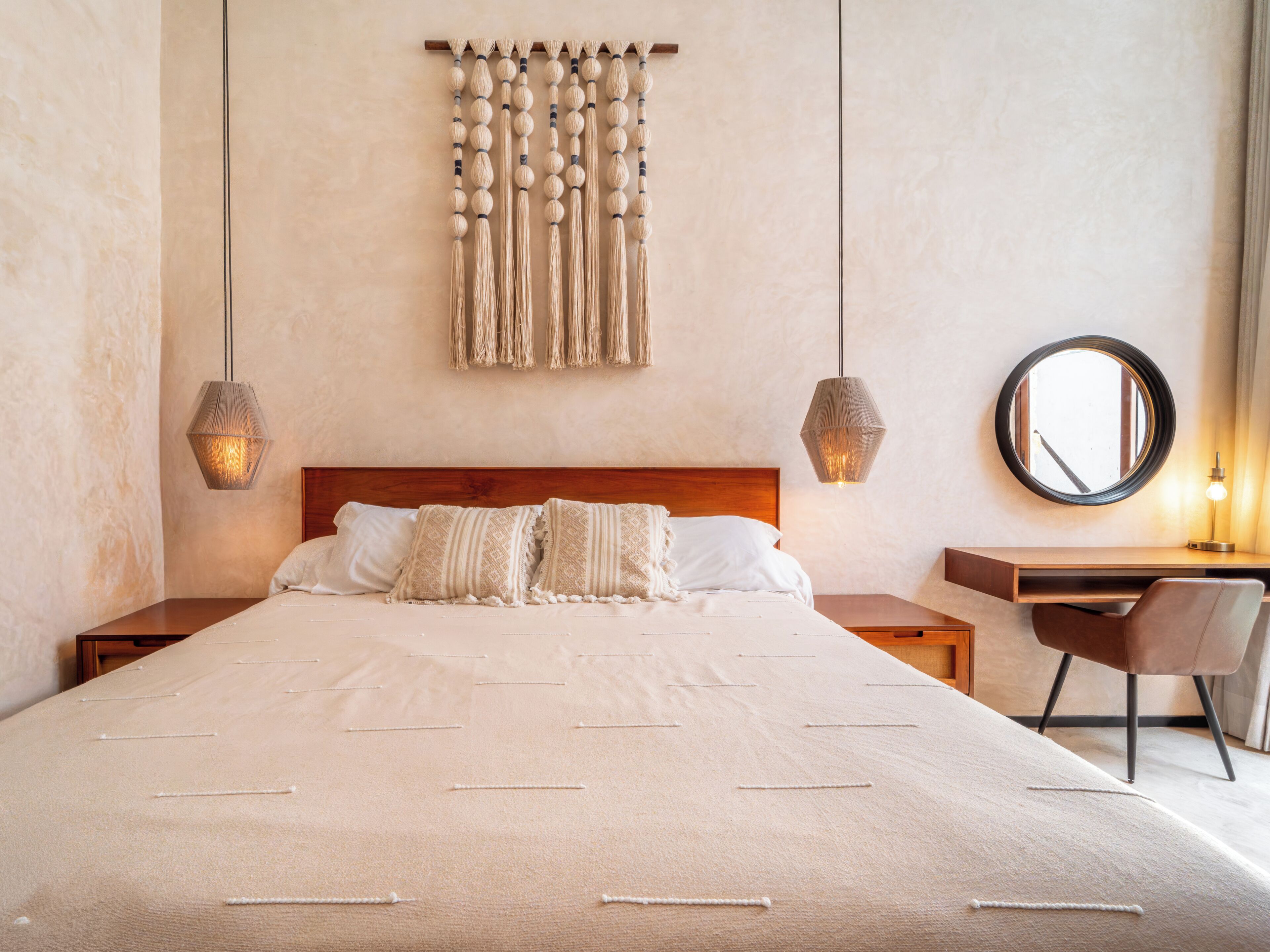 A bedroom with a large bed, two bedside tables, a desk with a chair, a round mirror, and a wall-mounted macrame piece.
