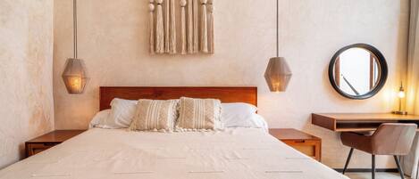 A bedroom with a large bed, two bedside tables, a desk with a chair, a round mirror, and a wall-mounted macrame piece.