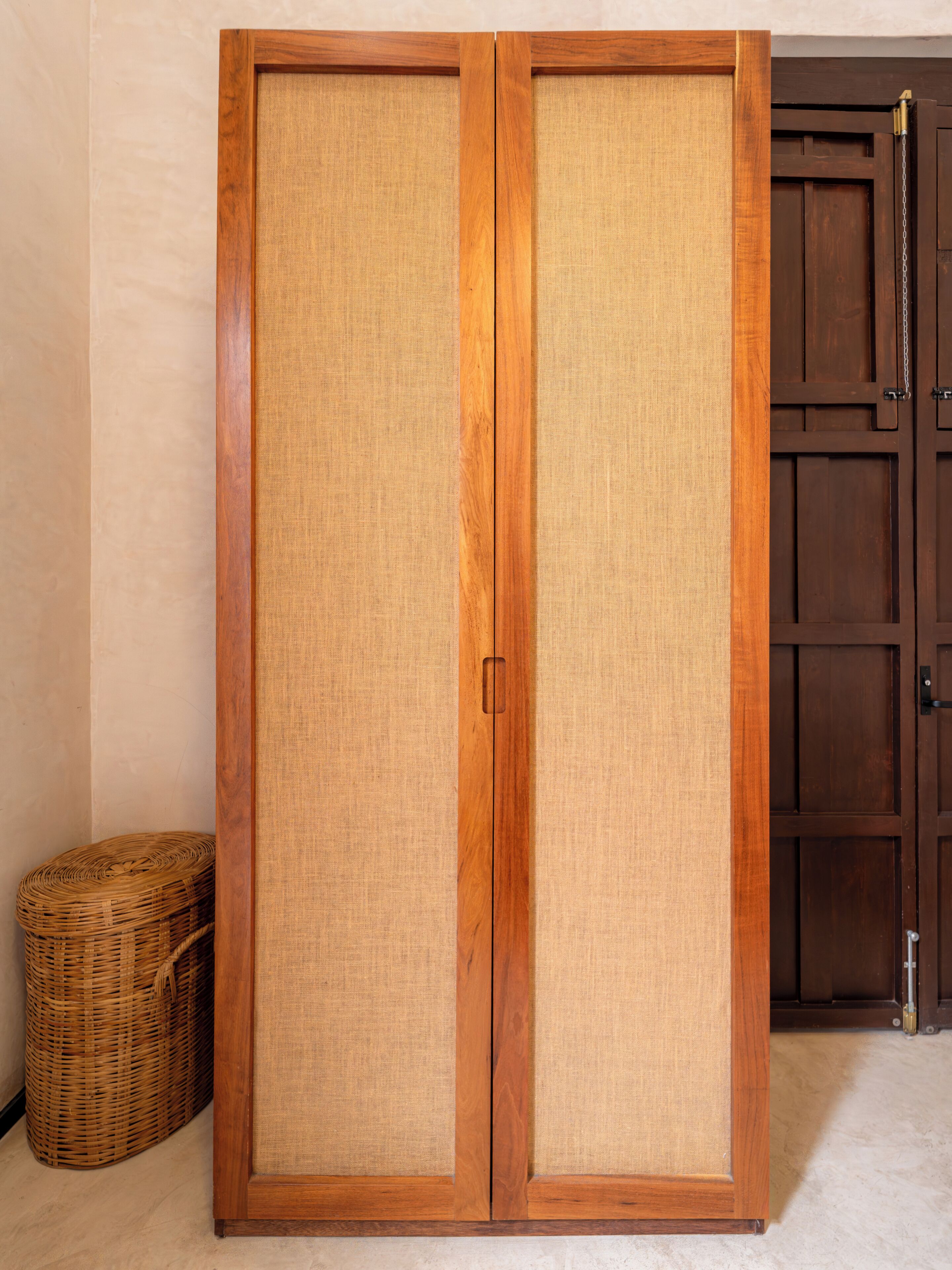 A wooden wardrobe with two doors, a wicker basket, and a wooden door in the background.