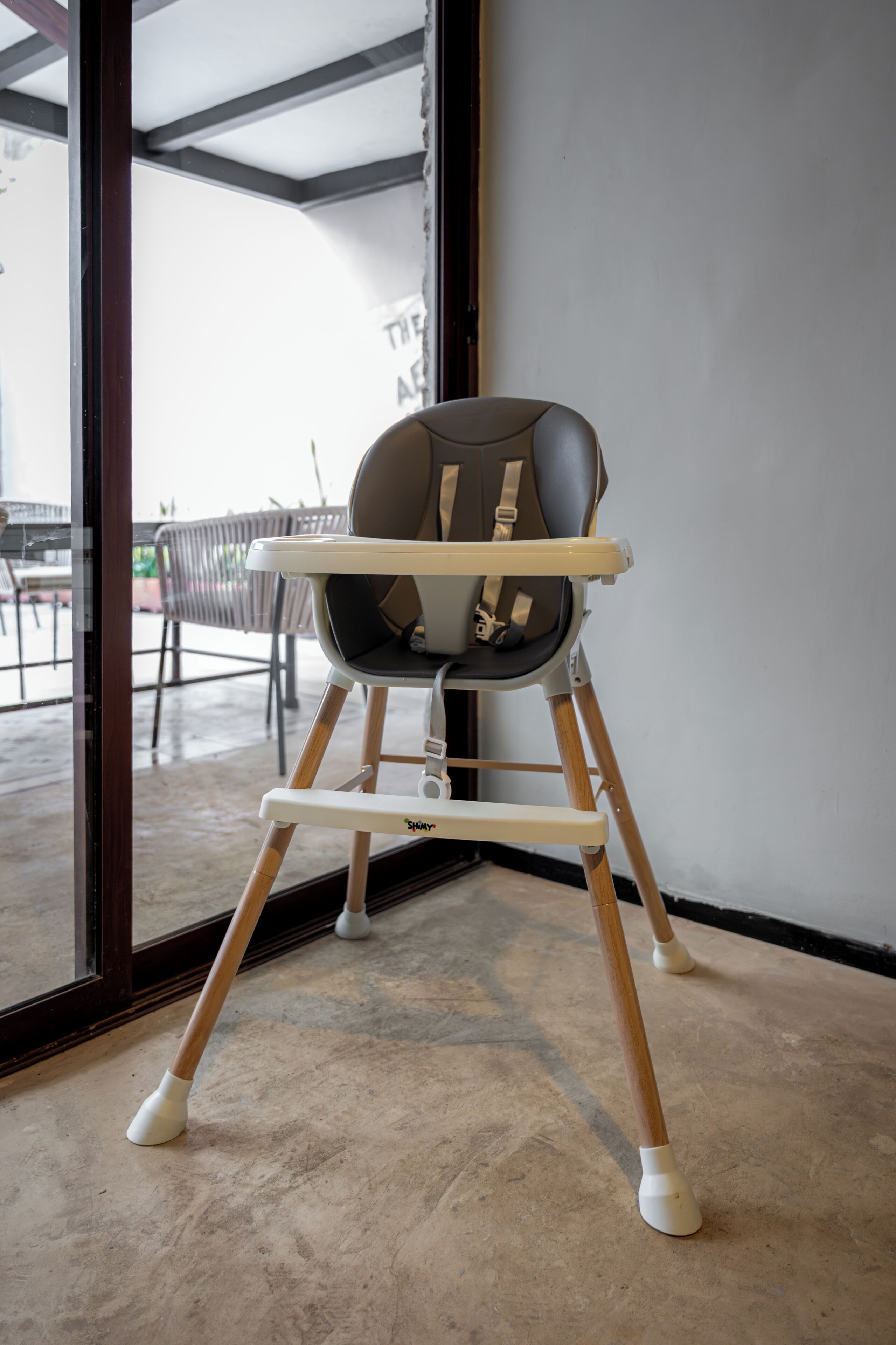 A high chair in a room with a large window, a concrete floor, and a visible outdoor area with chairs.