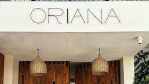En byggnadsentré med namnet ”ORIANA” ovanför dörrarna. Två hängande lampor av rotting hänger ovanför entrén.