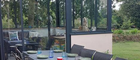 En uteservering med ett bord för fyra personer, omgiven av stolar, och en glasvägg med utsikt över trädgården.