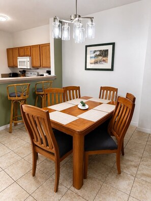 Un comedor con mesa y sillas de madera, una cocina al fondo y un cuadro en la pared.