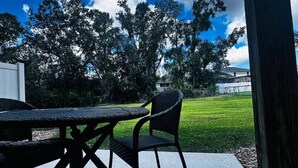 An outdoor patio with a table and chairs, a concrete floor, and a view of a grassy area with trees and a building in the background. - The Ideal Getaway V | 3BR near Disney & Pool (Kissimmee)