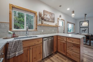A modern kitchen with wooden cabinets, a dishwasher, a large window, and a dining area with a table and chairs. - Unplug in Nature + Cabin in the Pines + Hot Tub, Game Room, Near the Lake (Ronald)