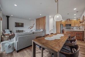 A modern kitchen with wooden cabinets, a central island, and a dining area with a wooden table and chairs. - Unplug in Nature + Cabin in the Pines + Hot Tub, Game Room, Near the Lake (Ronald)