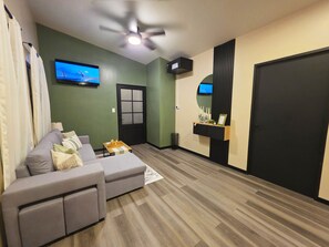 Uma sala de estar com um sofá cinza, um ventilador de teto, uma televisão, uma porta e uma pequena mesa com objetos decorativos.
