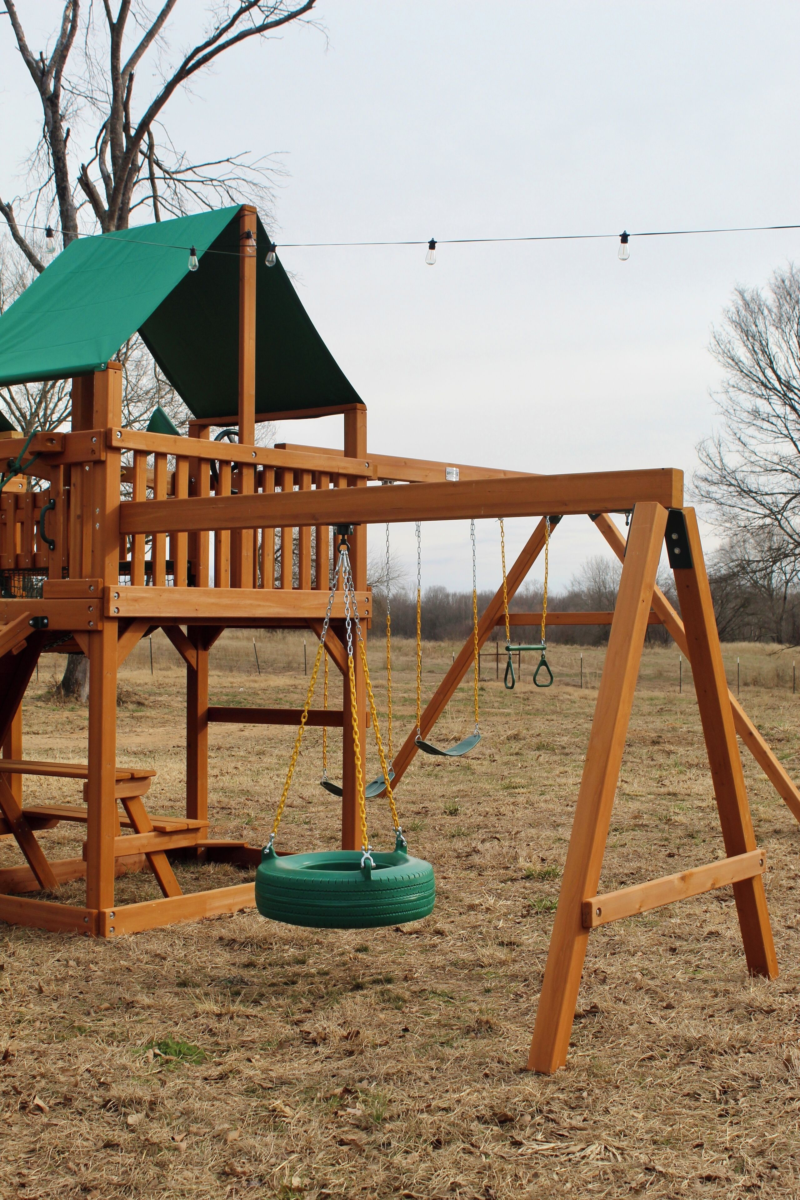 A wooden swing set with a green canopy, situated in a grassy area with leafless trees in the background.