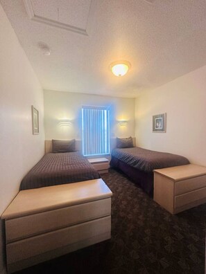 A hotel room with two single beds, a small bedside table, a dresser, and a window with blinds. - Your next adventure 3 (Kissimmee)