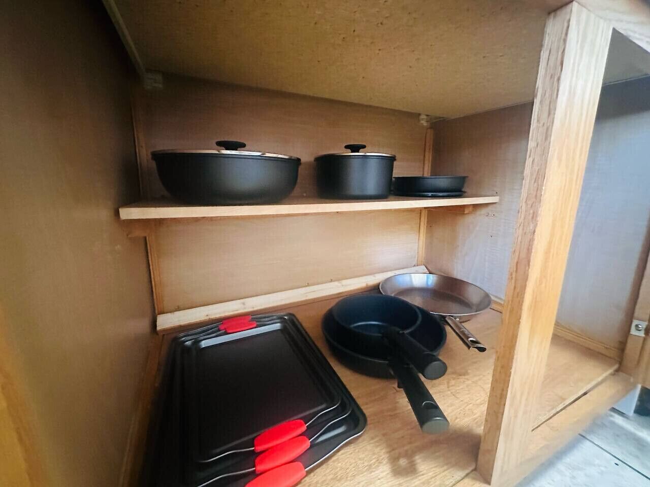 A compact kitchen with a wooden shelf holding black cookware, a frying pan, and a black tray with red handles.