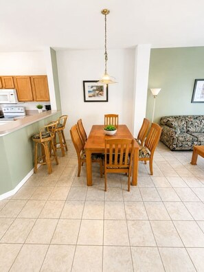 Een eethoek met een houten tafel en stoelen, een hanglamp, een keuken met houten kastjes en een groene bank.