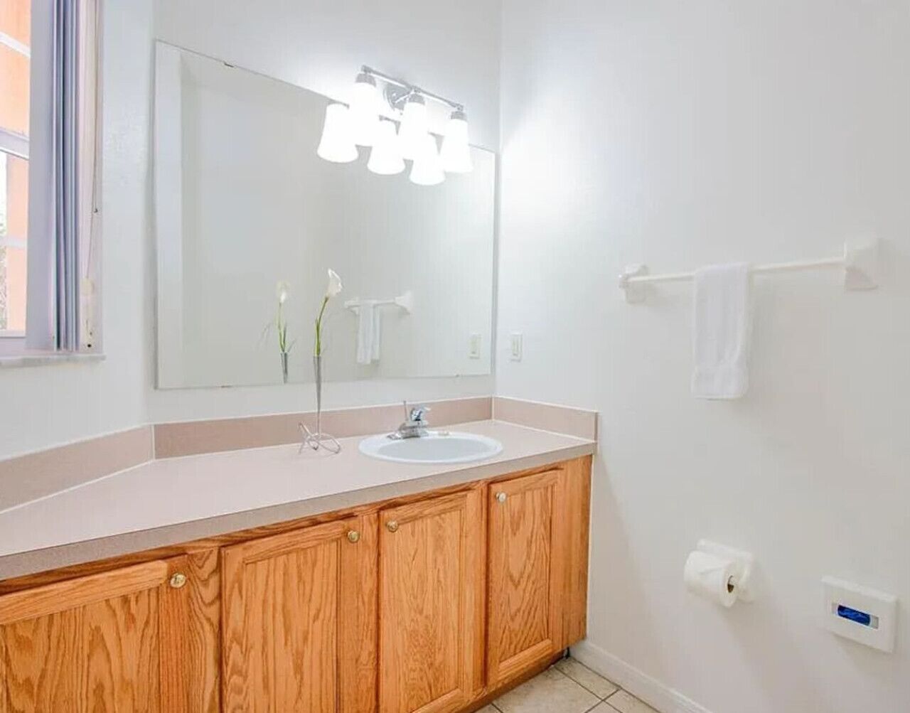 A bathroom with a wooden vanity, a white sink, a mirror with lights, a towel rack, and a toilet paper holder.
