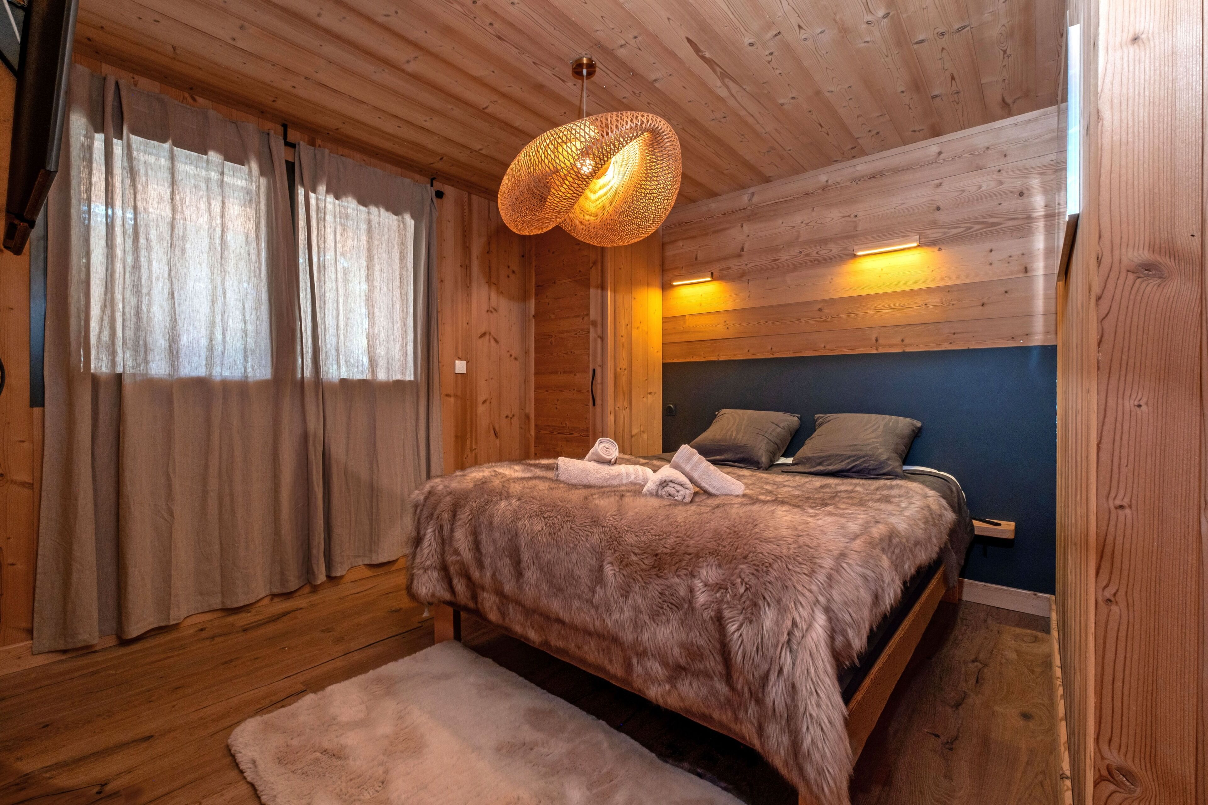 Une chambre dans une cabane en bois, avec un lit, des rideaux, un luminaire suspendu et un banc.