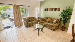 Un salon comprenant un ensemble de canapés, une table basse en verre, une plante en pot et une vue sur l’extérieur, visible à travers une porte vitrée.