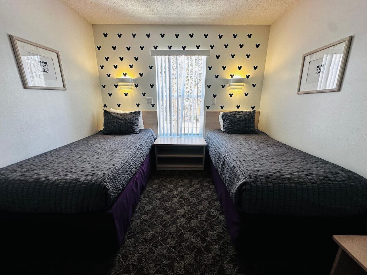 Une chambre avec deux lits simples, une fenêtre avec des rideaux, des lampes fixées au mur et des tableaux encadrés accrochés aux murs.