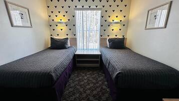 Une chambre avec deux lits simples, une fenêtre avec des rideaux, des lampes fixées au mur et des tableaux encadrés accrochés aux murs.