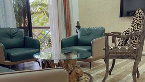 A living room with two green sofas, a wooden chair with a patterned cushion, a glass coffee table, and a TV mounted on the wall. - Comoé Lodge Hôtel Resort & Spa (Ekressinville)