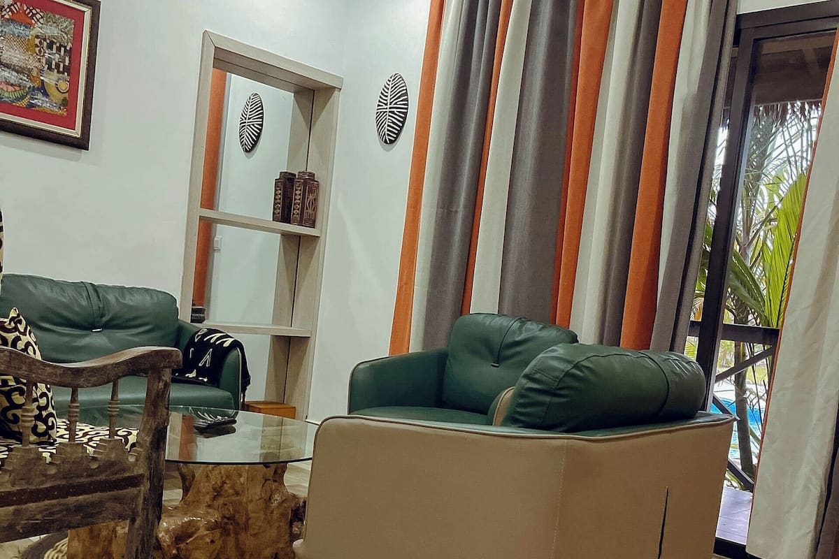 A living room with a green sofa, a wooden coffee table, and a patterned rug. There is a bookshelf with decorative items and a window with curtains.