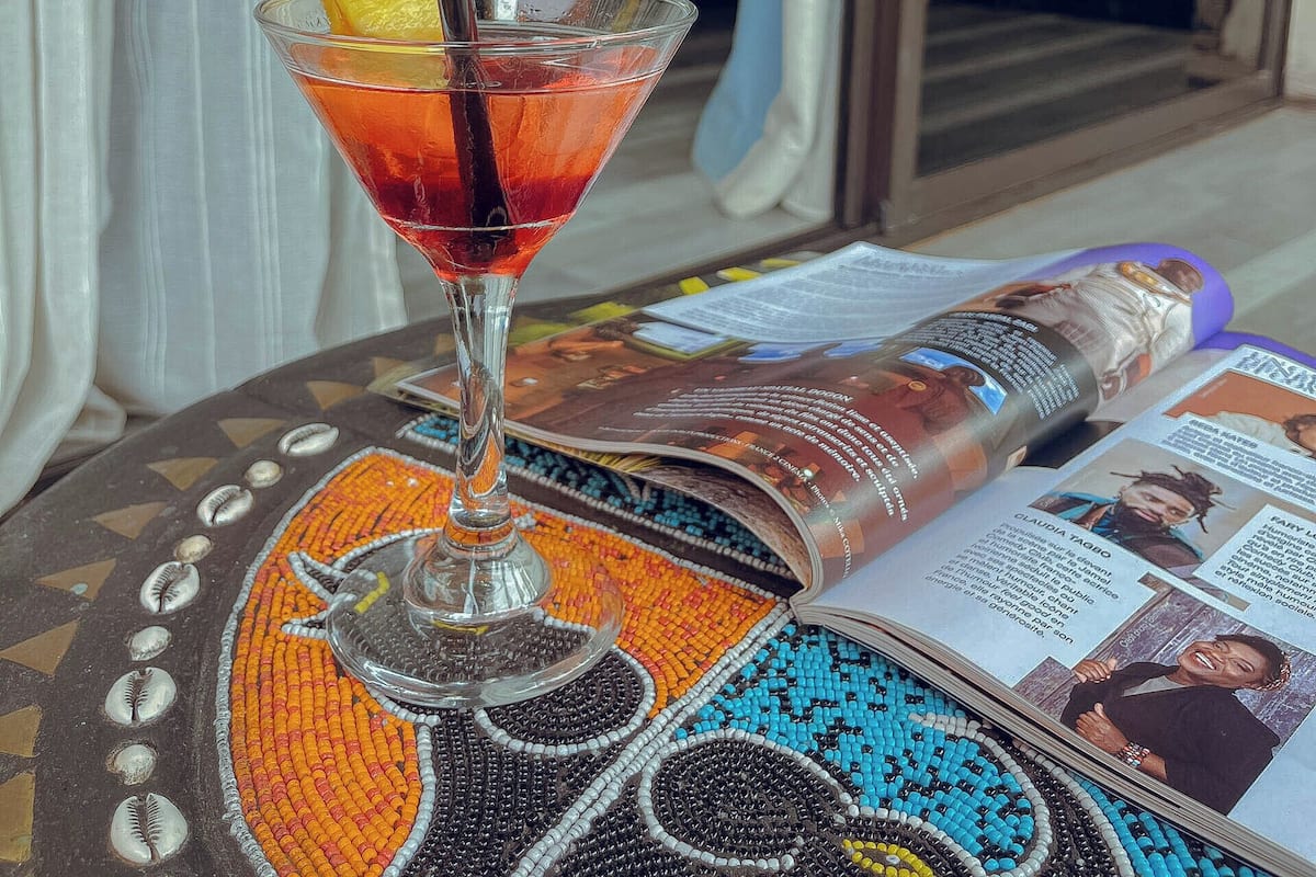 A martini glass with a lemon slice on a table with a magazine, in a room with a bed and a view of the outside.