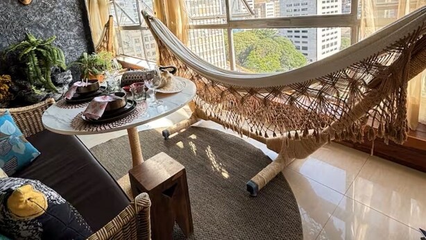 Een kamer met een groot raam, een hangmat, een ronde tafel met een glazen blad en rieten meubilair.