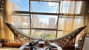Een kamer met een hangmat, een ronde eettafel gedekt met borden, glazen en een taart, en uitzicht op stadsgebouwen door grote ramen.
