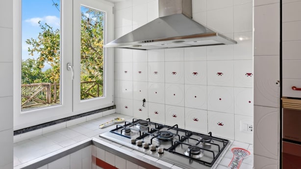 Una cocina con estufa a gas, azulejos blancos con diseños rojos, una ventana con vistas a árboles y una campana extractora de acero inoxidable.