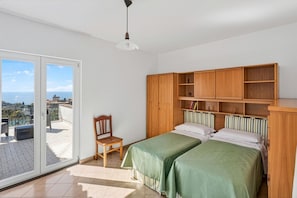 A bedroom with two beds, a wooden wardrobe, a chair, and a view of the city through a sliding door. - SorrentoVibes - The Lemon Grove Villa with private pool, garden and terrace (Massa Lubrense)