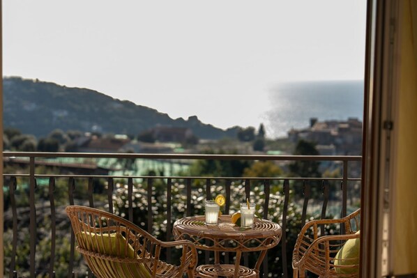 Ein Balkon mit Korbmöbeln, einem kleinen runden Tisch mit Getränken und einem herrlichen Blick auf das Meer und die Hügel.
