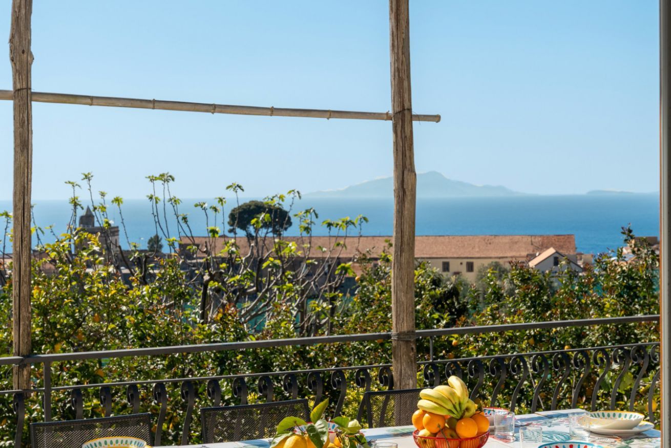 A balcony with a table set for dining, overlooking a coastal view with buildings and trees.
