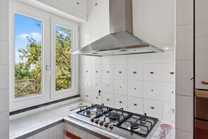 Una cocina con estufa a gas, azulejos blancos con diseños rojos, una ventana con vistas a árboles y una campana extractora de acero inoxidable.