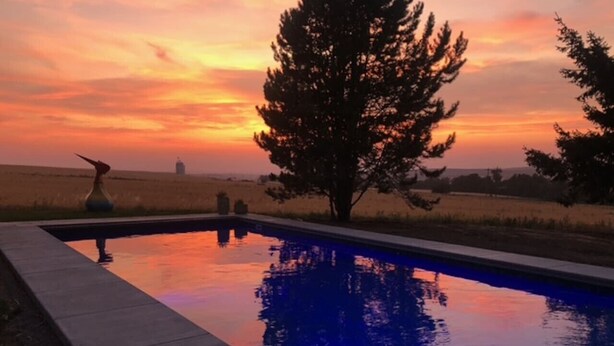 Une piscine avec vue sur le coucher de soleil, entourée d’arbres et donnant sur des champs ouverts.