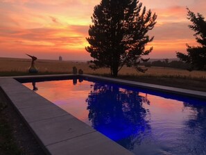Une piscine avec vue sur le coucher de soleil, entourée d’arbres et donnant sur des champs ouverts.