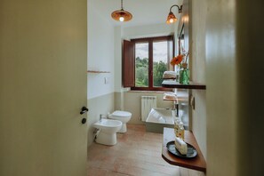 A bathroom with a toilet, bidet, and sink. There is a window with a view of greenery outside.