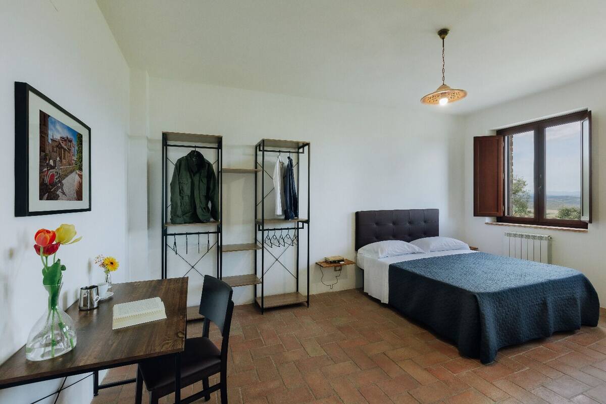 A bedroom with a bed, a desk with a chair, a shelf with clothes, a window, and a picture on the wall.