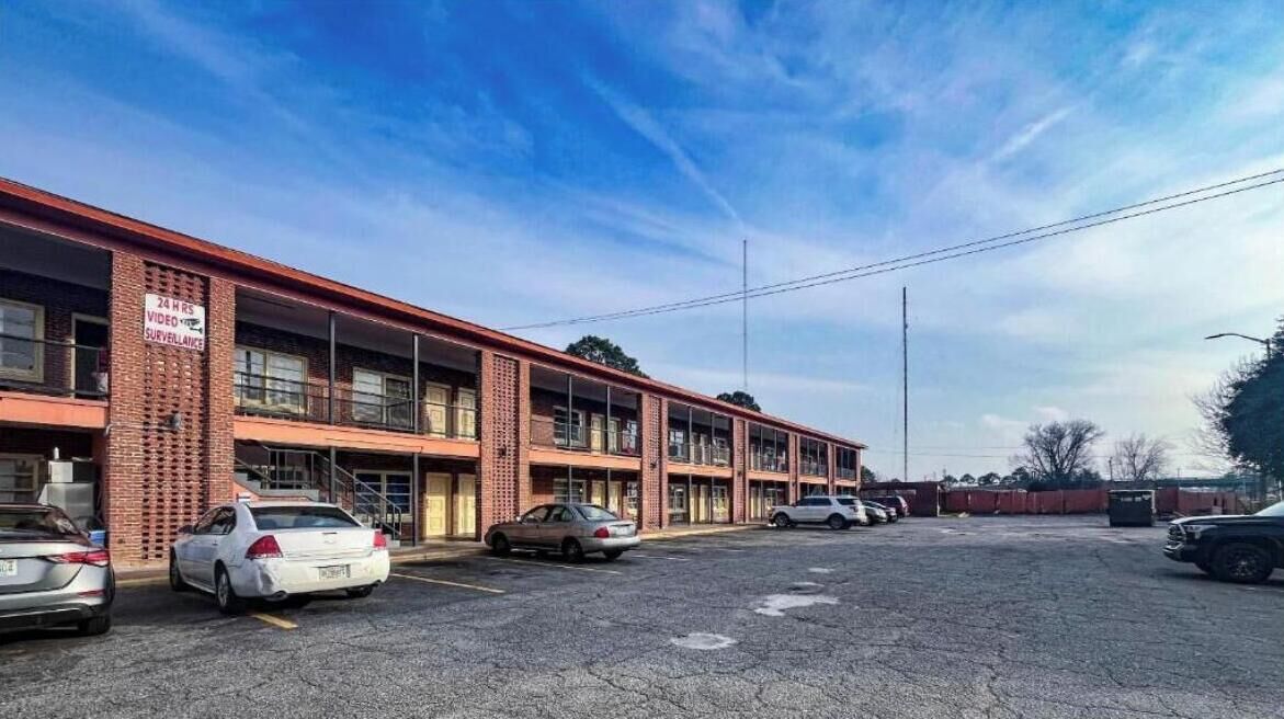 Un immeuble à plusieurs étages avec une façade en briques, un parking où sont garées plusieurs voitures et un ciel dégagé.
