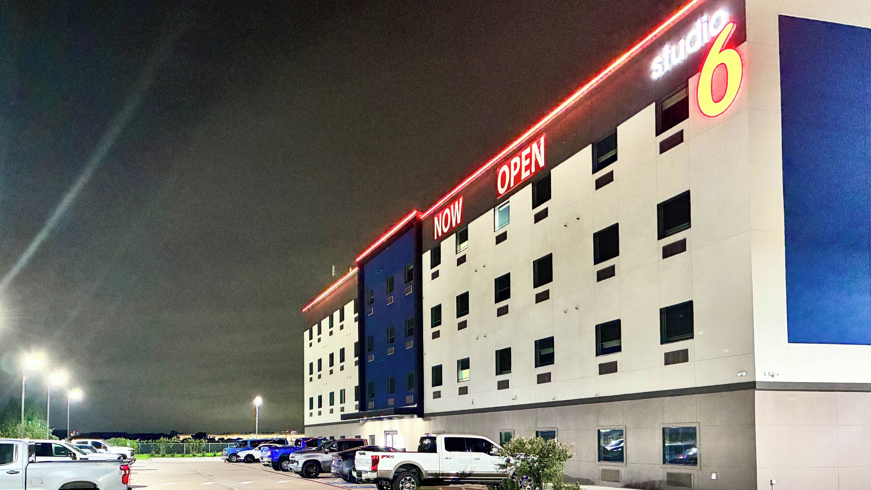 A multi-story hotel with a prominent 'OPEN NOW' sign, parking lot, and several parked vehicles.