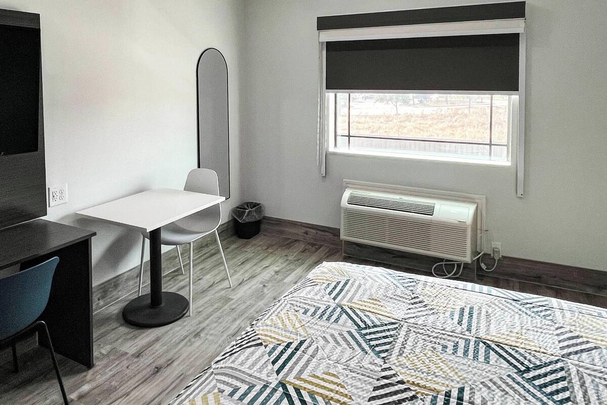 Una habitación pequeña y moderna con un escritorio, una silla y un espejo grande. Hay una ventana con vistas a un edificio.