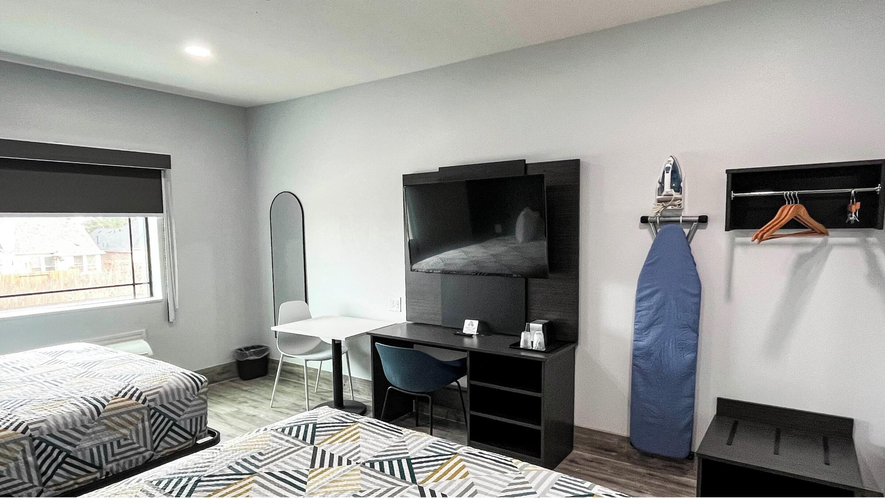 A modern hotel room with two beds, a desk, a chair, a TV, a mirror, and a hanging clothes rack.