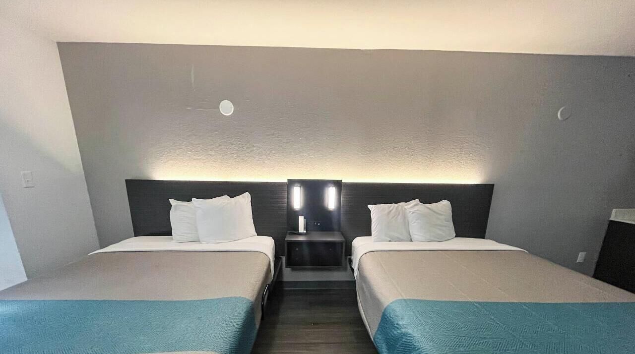 A hotel room with two beds, a nightstand with a lamp, and a wall-mounted light fixture.