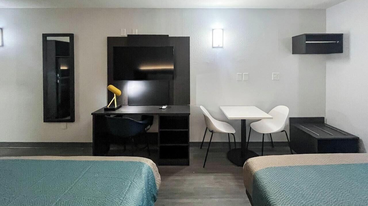 A compact hotel room with two single beds, a desk with a chair, a small table, and a mirror.