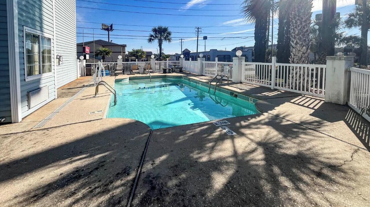 Une piscine rectangulaire, entourée d’une clôture blanche et dotée d’un escalier, ainsi que d’une terrasse en béton et de palmiers.