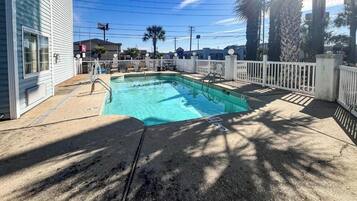 Une piscine rectangulaire, entourée d’une clôture blanche et dotée d’un escalier, ainsi que d’une terrasse en béton et de palmiers.