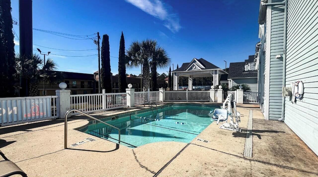 Una piscina con valla blanca, una zona de estar techada y un edificio al fondo.
