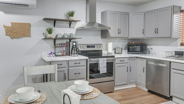 A modern kitchen with grey cabinets, stainless steel appliances, and a dining table set with white dishes. - Brand-New Sequim Escape: A/C, Fire Pit, Grill (Sequim)