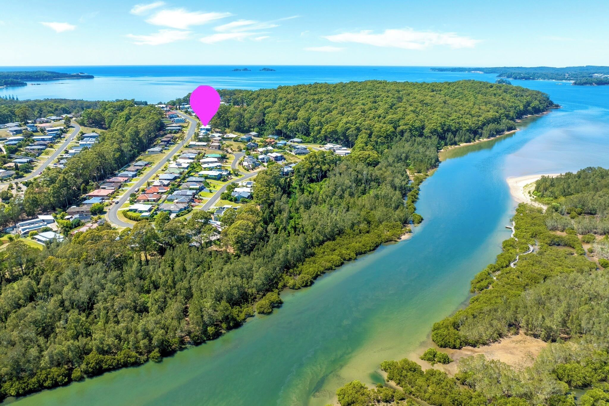 An aerial view of a coastal area with a river, dense forest, and a row of houses along the water's edge.