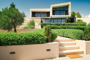 Una casa moderna de dos pisos con techo plano, ventanas grandes y un balcón. Hay escalones que conducen a la entrada, rodeados de setos bien cuidados.