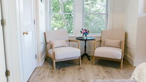 Une pièce avec deux fauteuils beiges, une petite table ronde ornée d’un vase de fleurs et une vue sur les arbres, visible à travers de grandes fenêtres.