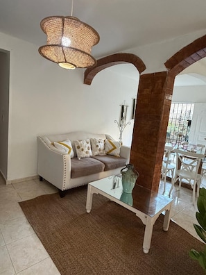 Una sala de estar con sofá blanco, una mesa de centro de vidrio, una lámpara colgante de mimbre y vista al área de comedor a través de un arco.