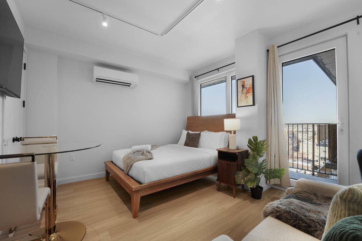 Quarto moderno com cama de madeira, mesa de cabeceira com abajur, planta em vaso e vista da cidade por uma janela grande.
